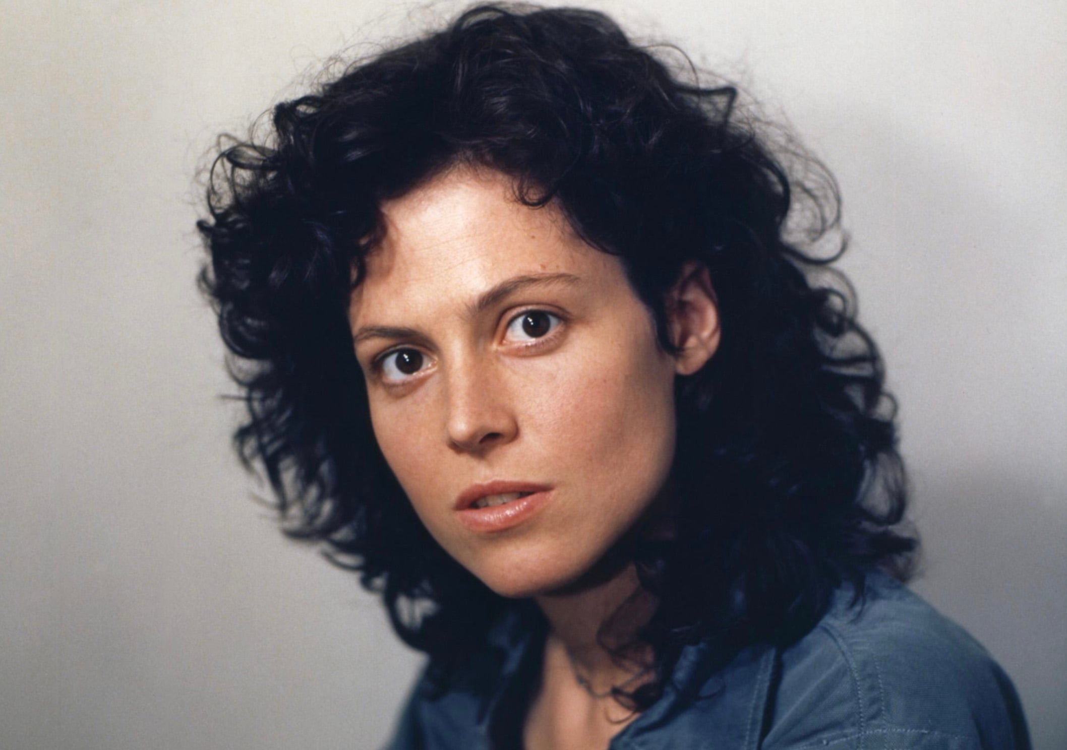A woman with curly dark hair and a calm expression looks directly at the camera, wearing a blue shirt against a plain light background.