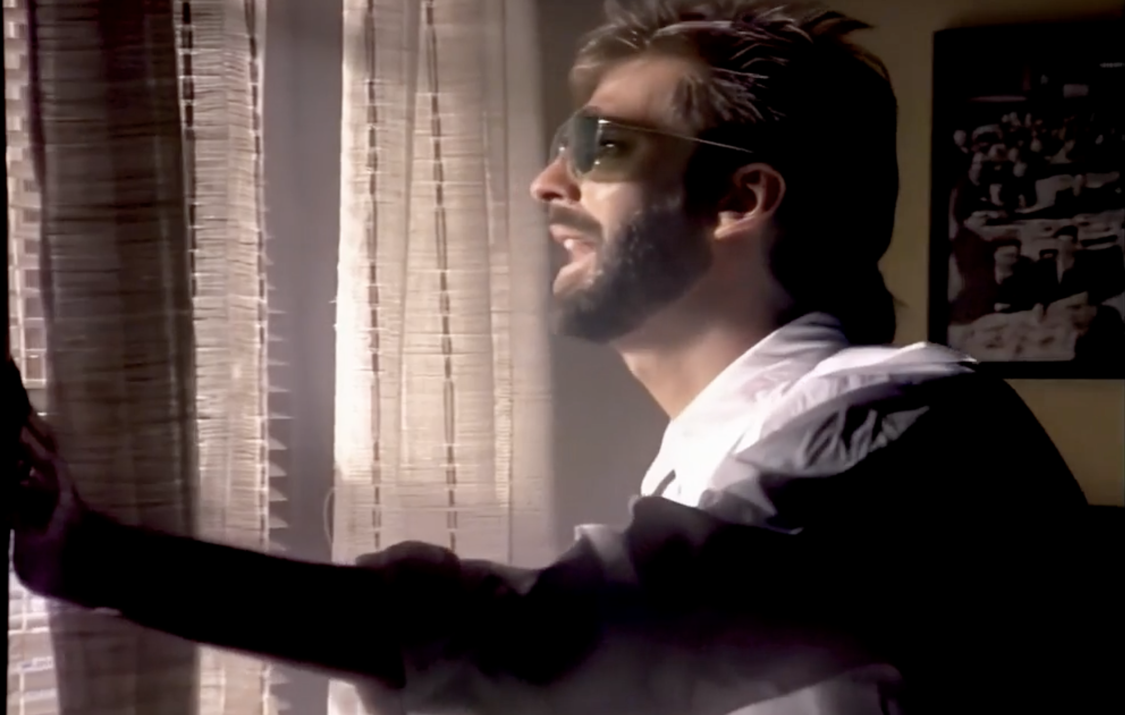 A man with a beard and sunglasses, wearing a white shirt, stands beside a window with sheer curtains, looking outside with his mouth slightly open, as light filters into the room.