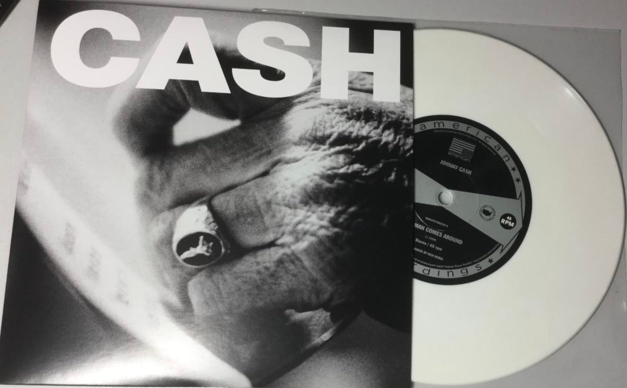 A black-and-white album cover with a close-up of a hand wearing a ring labeled "CASH," partially covering a white vinyl record. The record's label reads "American Recordings.