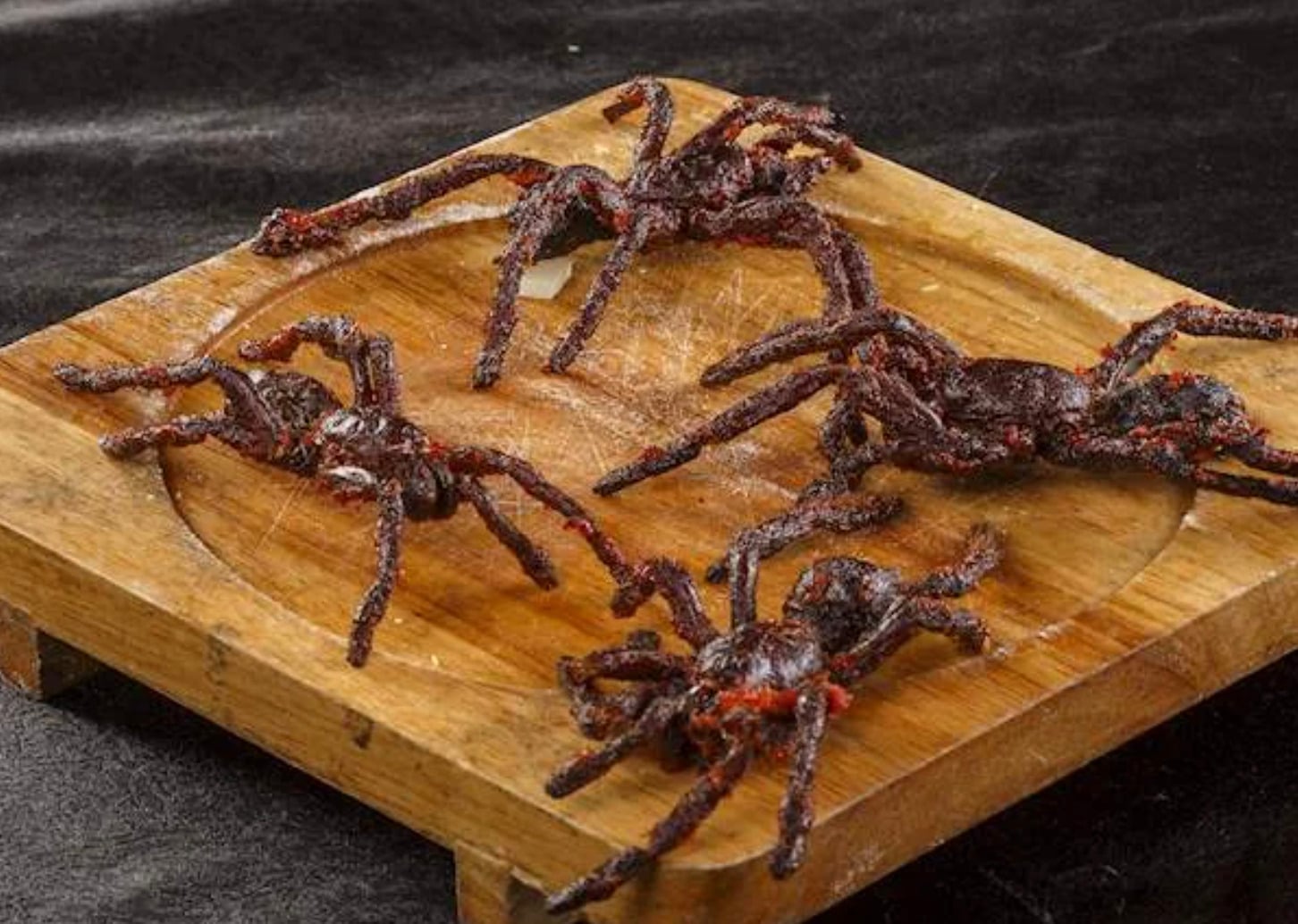 Four dark, crispy fried tarantulas are arranged on a wooden serving tray with a circular indentation, set against a black background.
