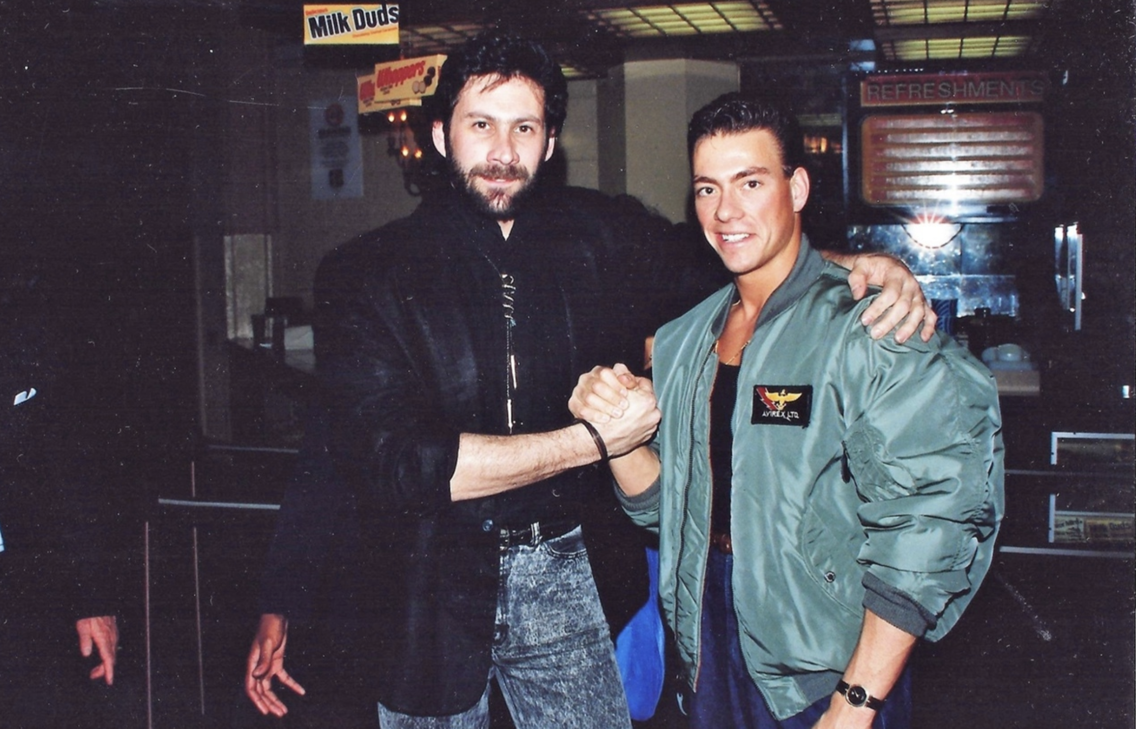 Two men pose together indoors, smiling and clasping hands. One wears a dark jacket and jeans, the other wears a green bomber jacket with a patch, and there is a Milk Duds sign visible in the background.