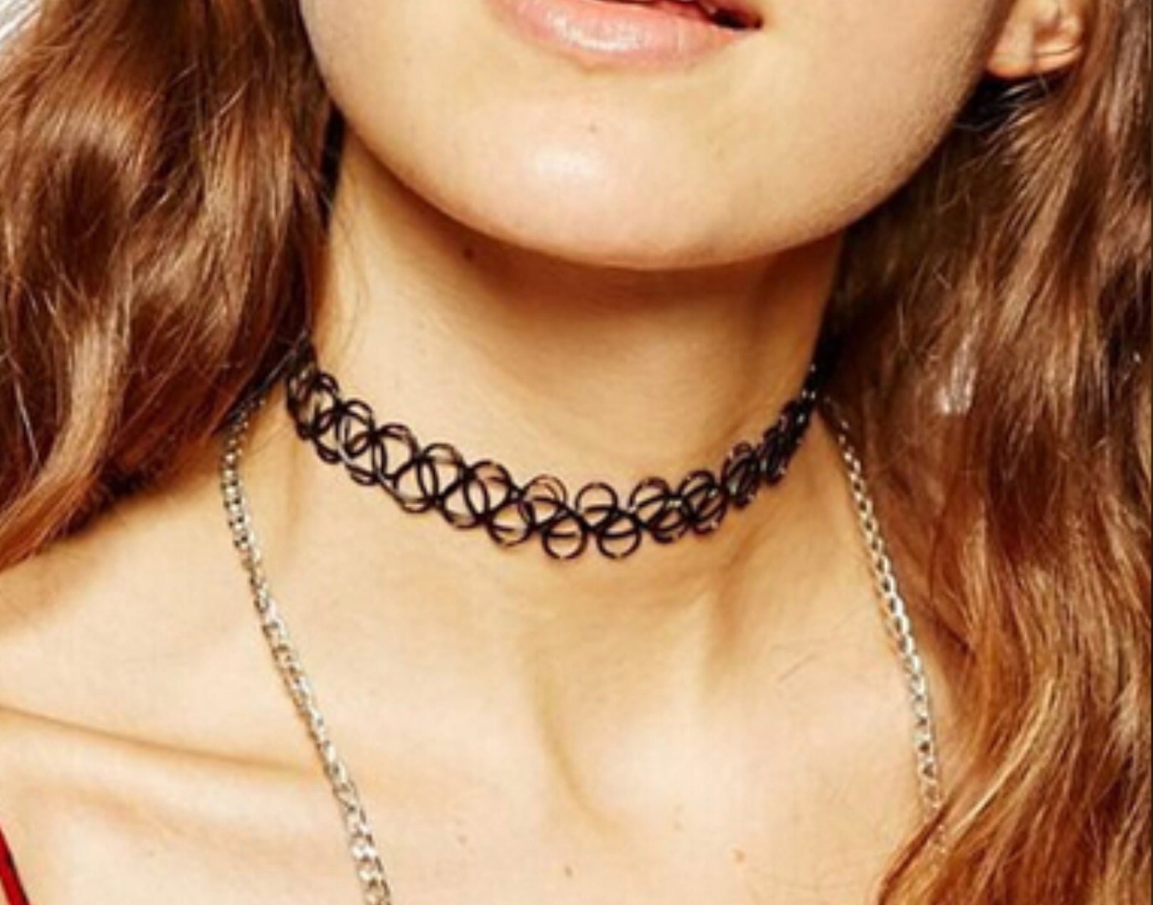 A person with wavy brown hair wears a black tattoo choker necklace and a silver chain necklace. The lower half of their face and neck are visible.