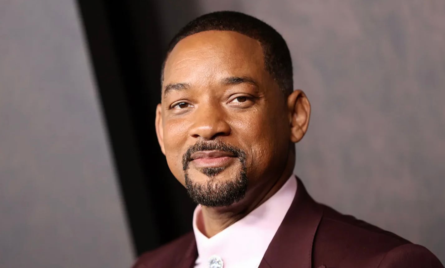 A man with short hair, a trimmed beard, and mustache, wearing a maroon suit jacket and pale pink shirt, smiles gently while looking at the camera against a blurred gray background.