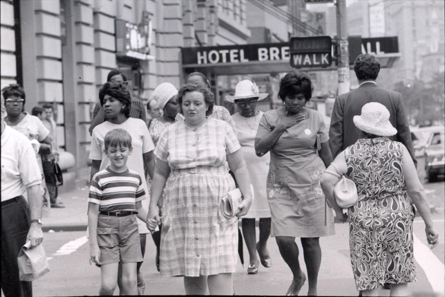 A black-and-white photo shows a busy city sidewalk; people of various ages and dress are walking. A “HOTEL BRE” sign and “WALK” signal appear overhead. Some pedestrians look ahead, others engage with each other.