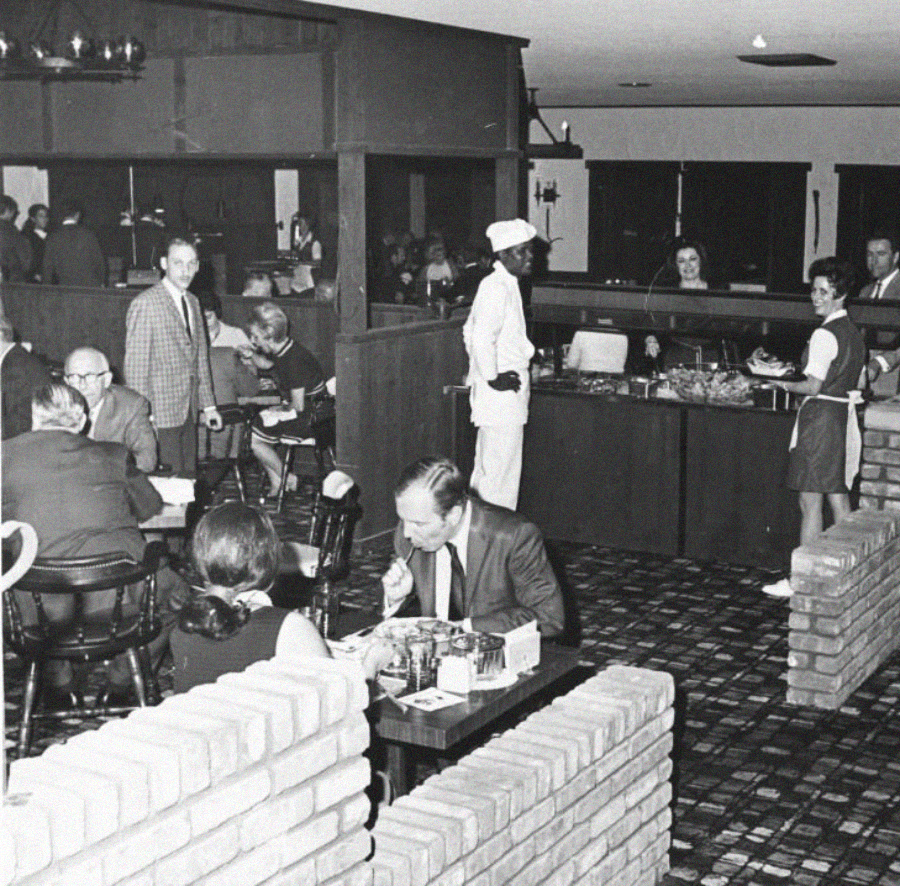 Black-and-white photo of a busy restaurant with diners seated at tables, several people serving food at a buffet, and staff dressed in uniforms. The decor includes brick partitions and patterned carpeting.