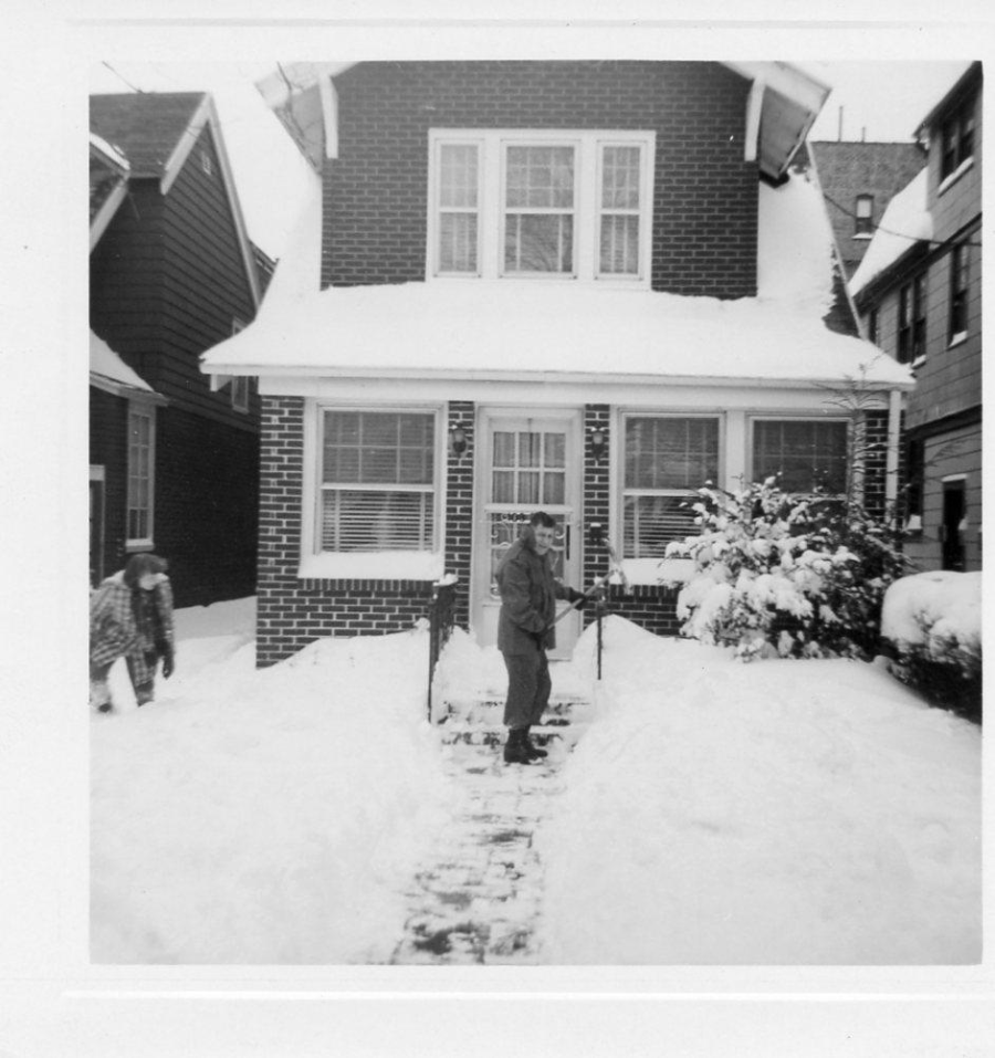 A person shovels snow on the walkway in front of a brick house, while another person walks through deep snow on the left side. Snow covers the ground, roof, and bushes in a residential neighborhood.