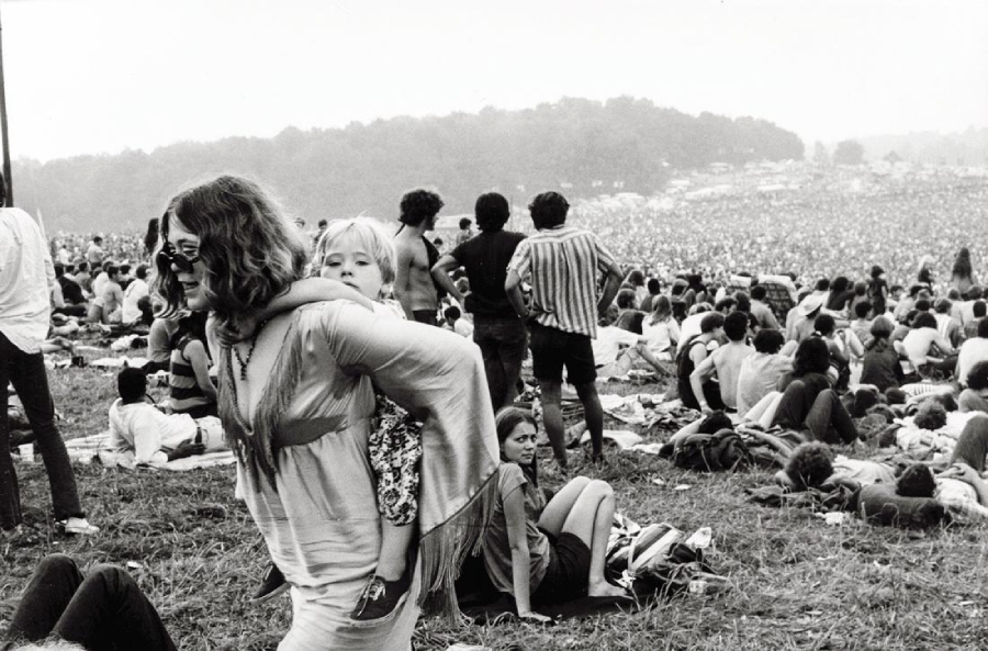 A black-and-white photo of a large outdoor music festival crowd. In the foreground, a woman in sunglasses carries a child on her back. People sit and relax on the grassy hillside, with thousands visible in the background.