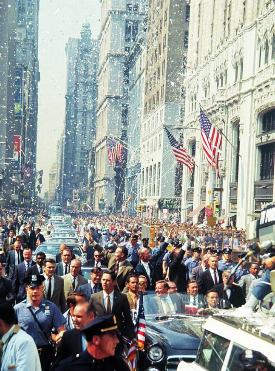 A large crowd lines a city street with American flags on buildings, confetti falling, police officers and cars leading a procession, as people watch and cheer during a parade in a bustling urban setting.