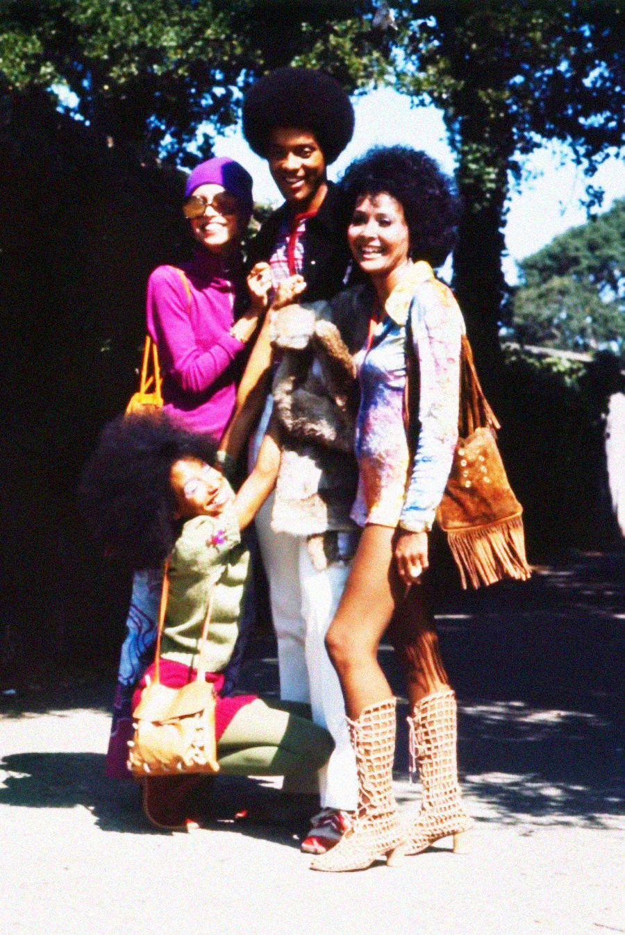 Four people pose outdoors in bright 1970s clothing, including patterned outfits, fringe purses, platform sandals, and Afro hairstyles. They are smiling and appear happy, with trees and sunlight in the background.