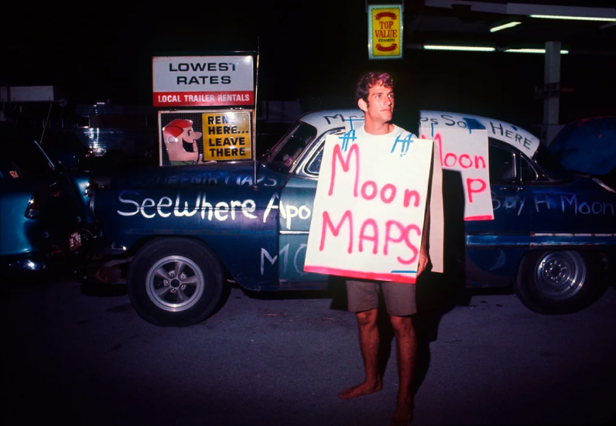 A man stands barefoot at night wearing a large sandwich board sign that reads "Moon MAPS" in red letters. He is in front of a vintage car with words written on its side and signs for trailer rentals visible in the background.