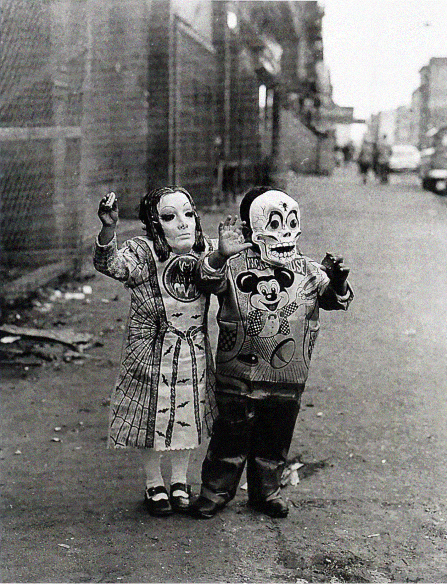 Two children stand on a city sidewalk wearing full masks and costumes; one wears a skeleton mask and Mickey Mouse sweater, the other a doll-like mask and plaid dress. The background is slightly blurred, showing buildings and a parked car.