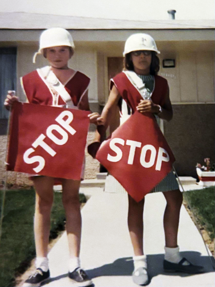 Two children wearing white helmets and red sashes hold large red stop signs while standing on a sidewalk in front of a house. It is sunny, and both appear to be school crossing guards.