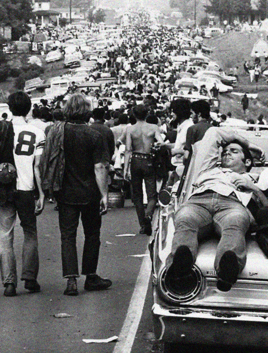 A black-and-white photo shows a large crowd walking on a road filled with parked cars. In the foreground, a man lounges on the trunk of a car while others walk past him. The atmosphere appears busy and relaxed.