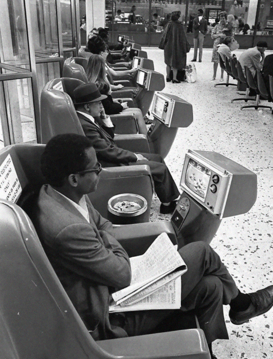 People sit in rows of retro lounge chairs at an airport, each chair equipped with a small television. Some people read newspapers while others watch TV. The setting appears busy, with travelers and staff in the background.