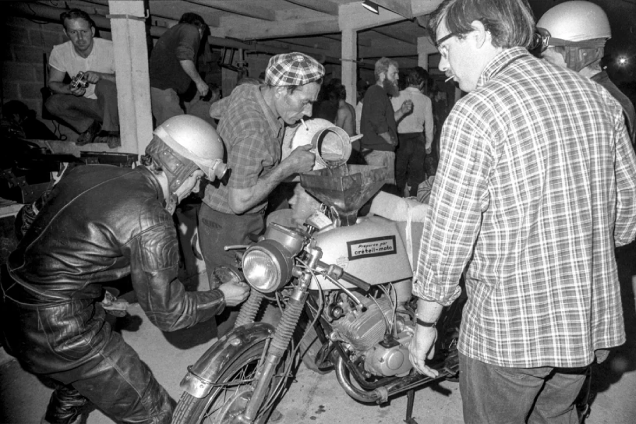 A group of men gather around a motorcycle in a garage, some wearing helmets and leather gear, while another pours liquid into the bike’s tank. Others watch or work nearby under fluorescent lights. The scene is busy and focused.