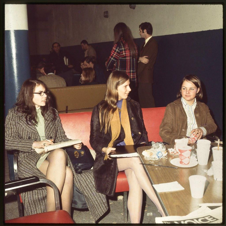 Three women sit on a red couch with papers, drinks, and food on the table in front. Other people sit and stand in the background, conversing in a room with blue and white walls. The scene has a 1970s vintage style.