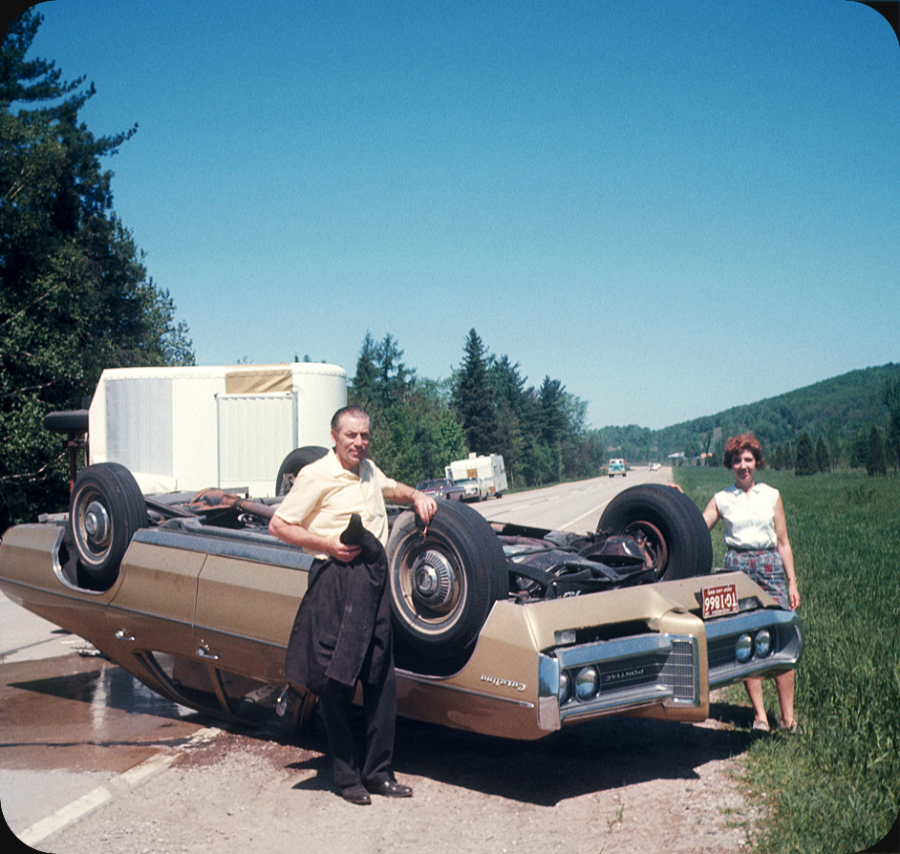 A man and woman stand next to an overturned vintage car on the side of a rural road, with a trailer and trees in the background on a sunny day.