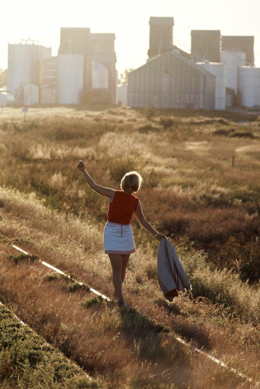 A person in a red top and white skirt walks barefoot along a railway track through tall grass, holding a jacket, with grain elevators and silos visible in the background.