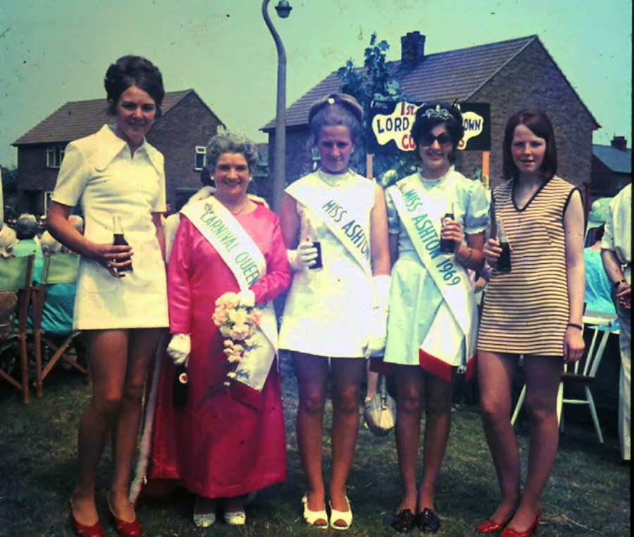 Five women pose outdoors at a community event. Three wear sashes labeled "Carnival Queen" and "Miss Ashton." They are dressed in 1960s-style outfits, with houses and a sign in the background.
