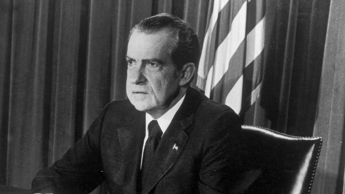 A man in a suit sits at a desk in front of a curtain and part of an American flag, looking serious. The photo is black and white.