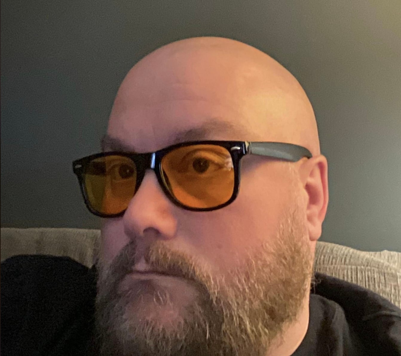 A bearded person with a shaved head is wearing black-framed glasses with amber-tinted lenses, sitting indoors against a neutral-colored wall.