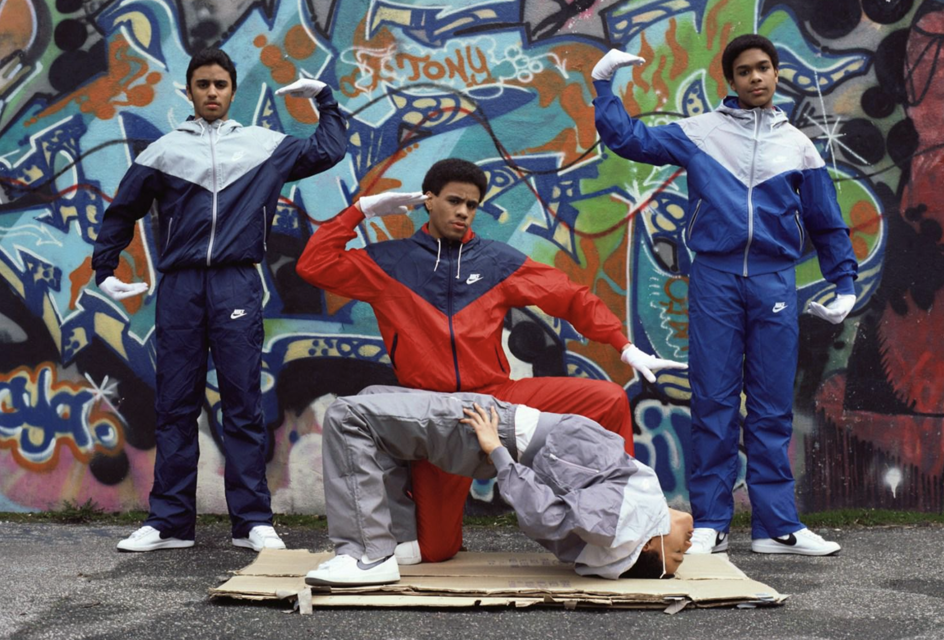 Four people in colorful tracksuits and white gloves pose in front of a graffiti wall. Three stand upright with arms raised, while one performs a backbend on cardboard in front of them.