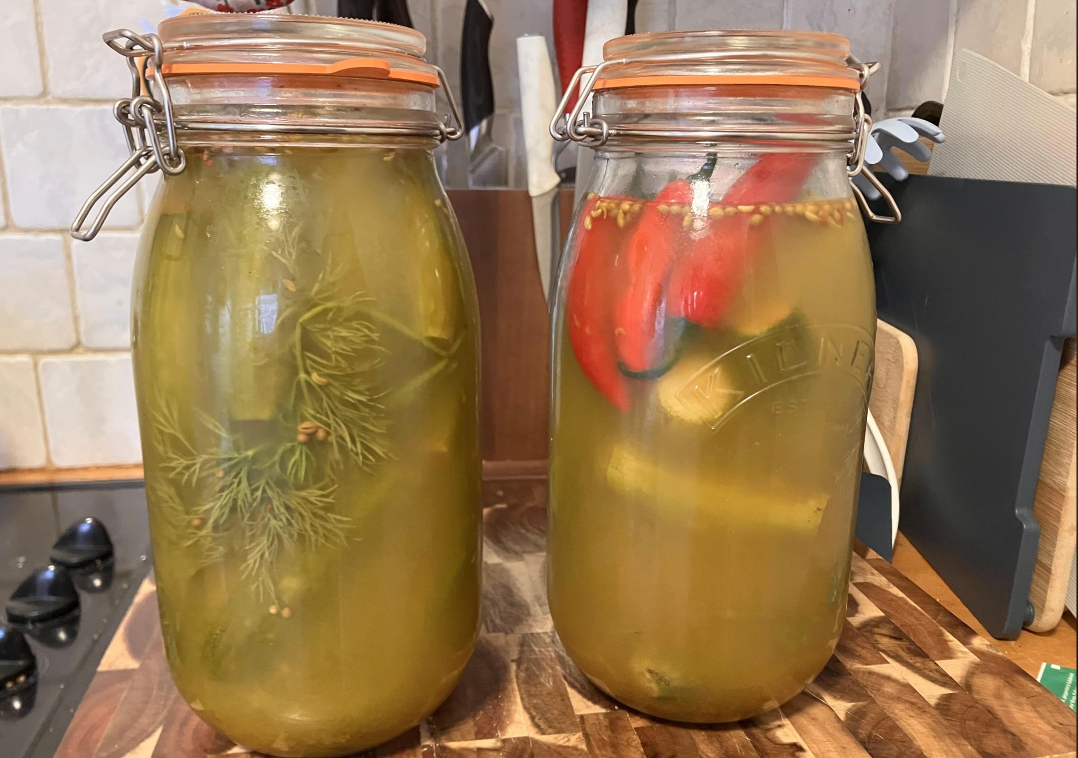 Two glass jars filled with homemade pickles sit on a wooden countertop. One jar contains dill, and the other has red peppers and spices. Both jars have metal clip-top lids and are filled with brine.