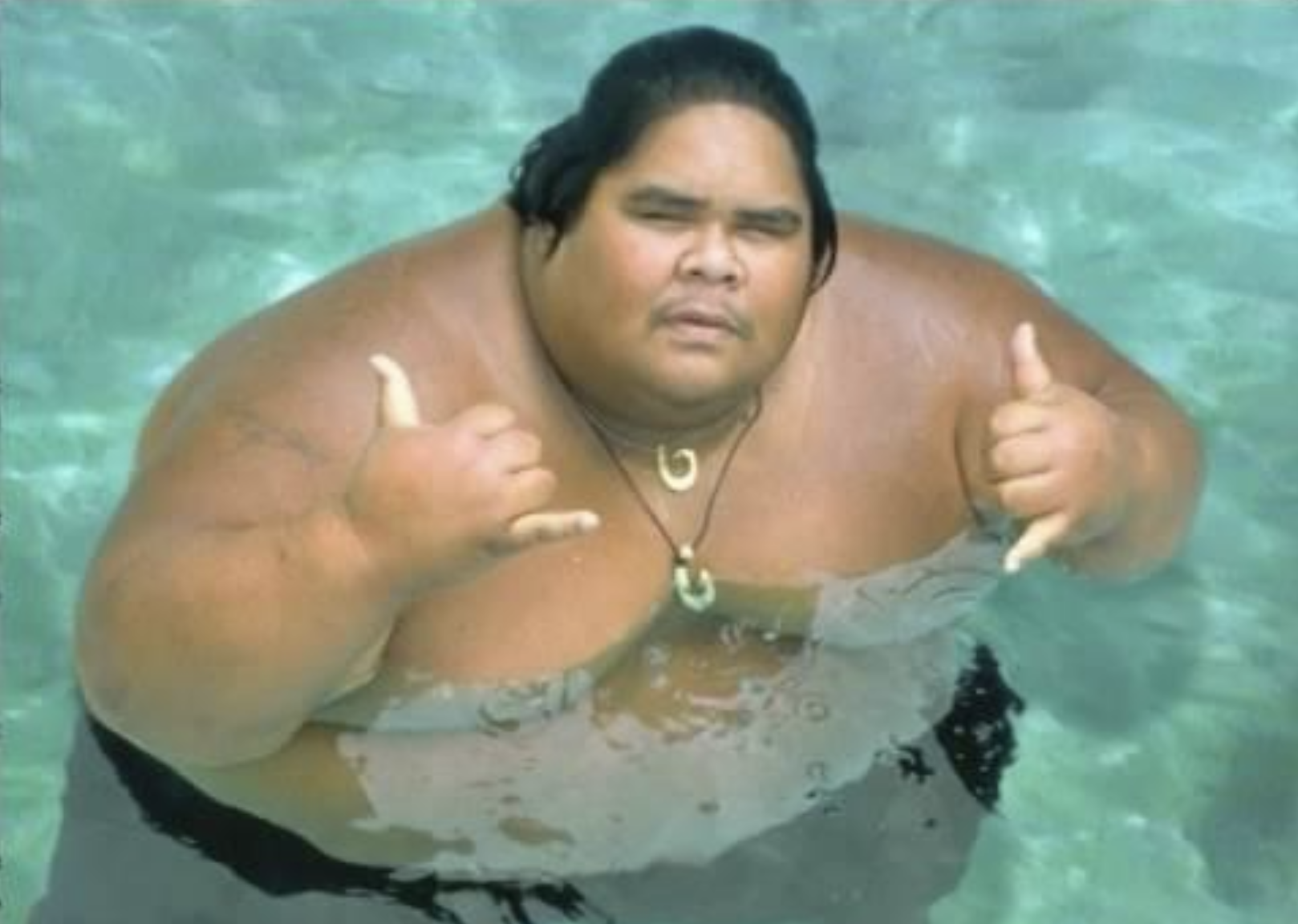 A large man with long dark hair stands in a pool of clear water, making a shaka hand gesture with both hands. He wears a necklace and looks directly at the camera.