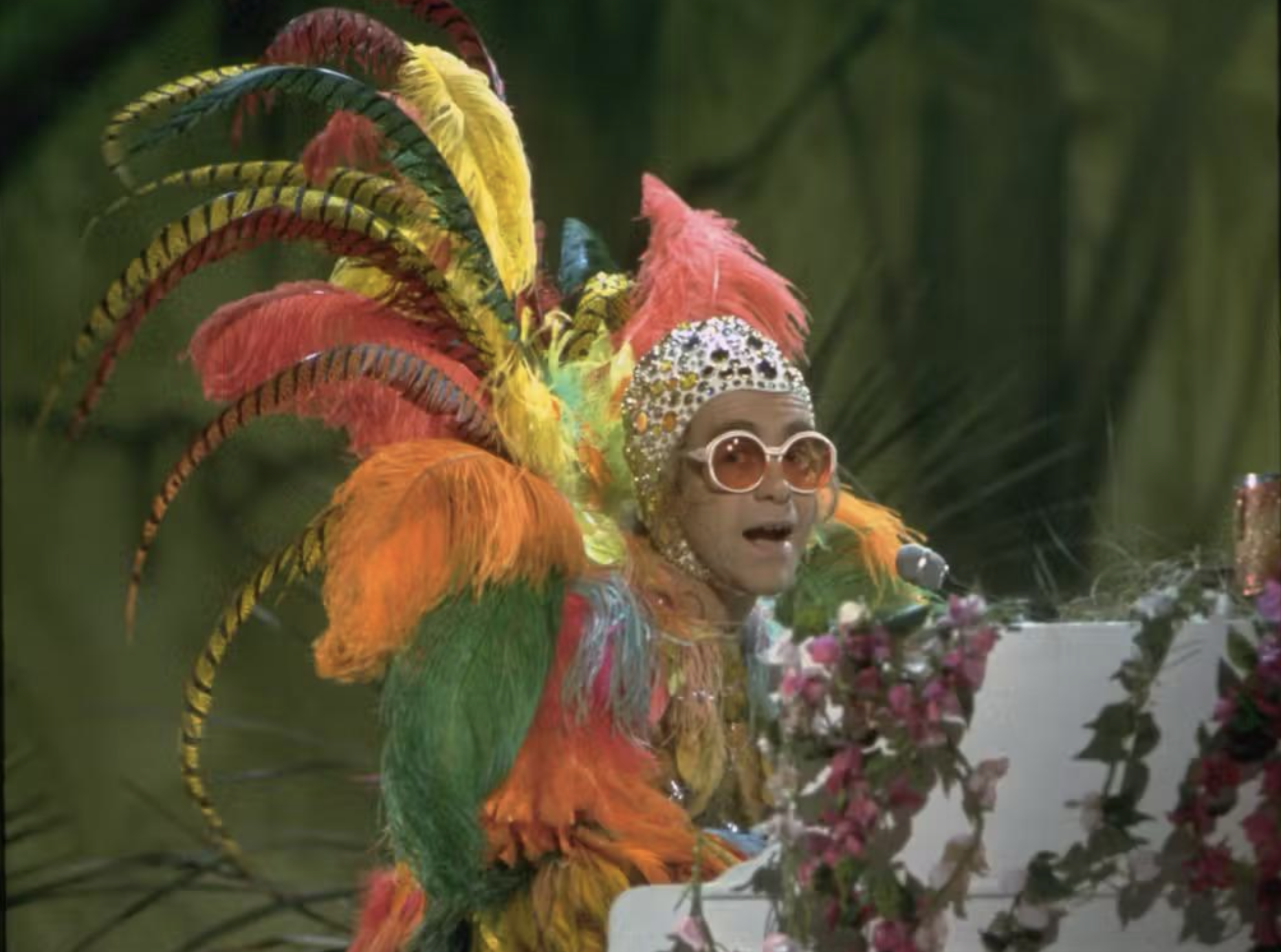 A person in large, colorful feathered costume and round glasses plays a white piano decorated with flowers, set against a green background.