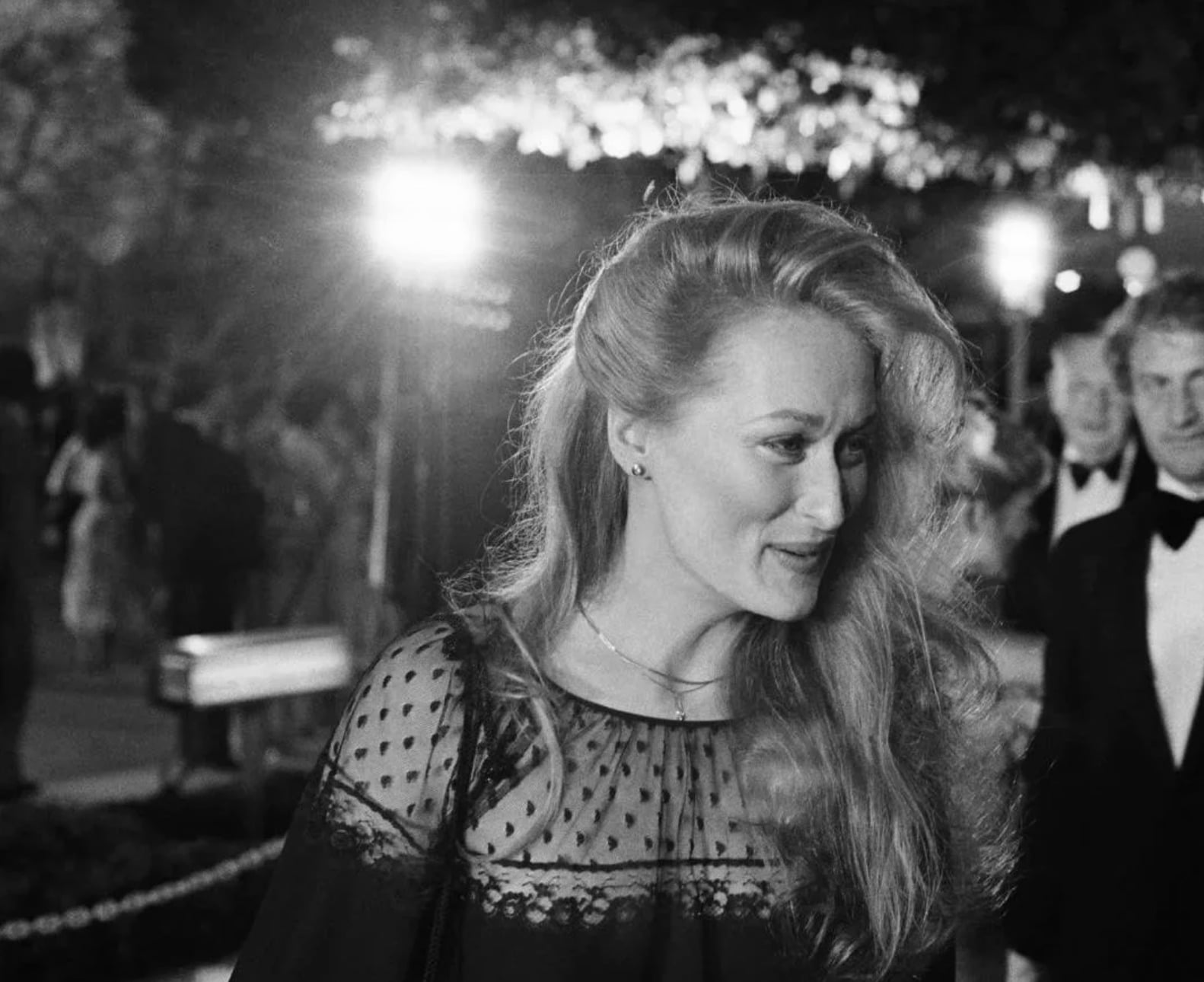A woman with long, wavy hair in a sheer, polka-dot dress attends a formal event at night. She is outdoors with people and bright lights in the background. The image is in black and white.