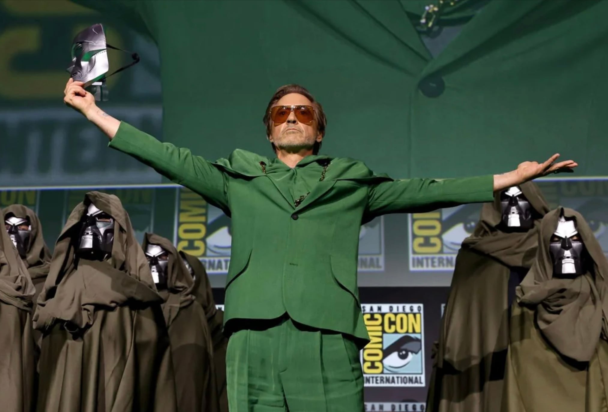 A man in a green suit stands on stage with arms outstretched, holding a metallic mask. He is surrounded by people wearing hooded cloaks and similar masks. Comic-Con International signs are visible in the background.