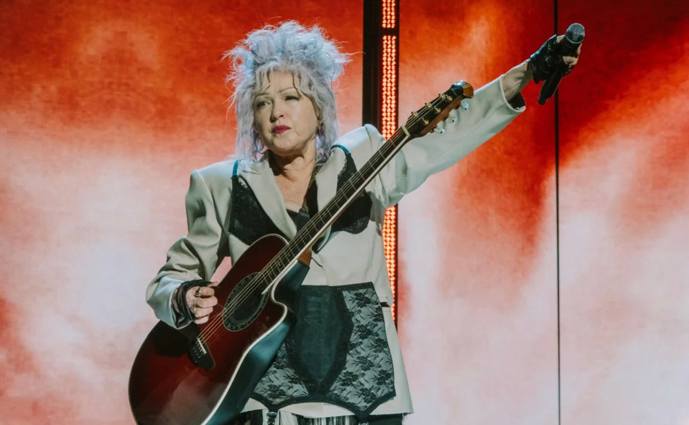 A woman with spiky gray hair holds a guitar and raises her arm while performing on stage, wearing a light-colored suit with dark accents and fingerless gloves, against a red and white lit backdrop.