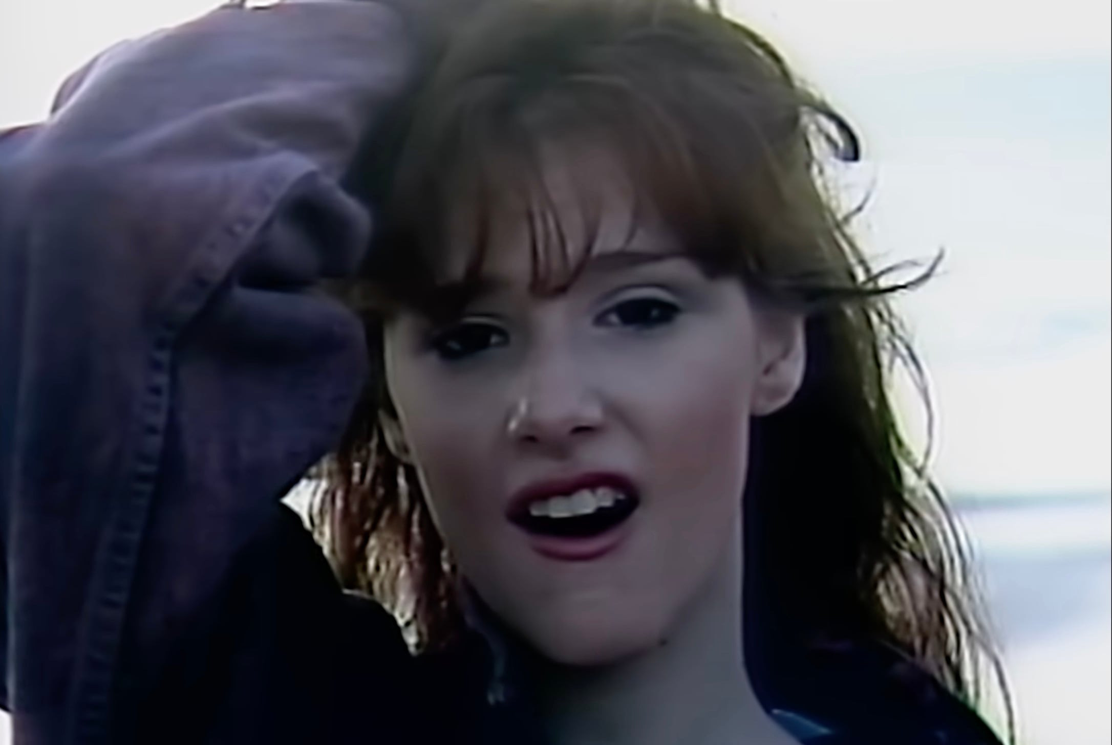 A young person with fair skin and long brown hair stands outdoors, wearing a loose jacket and touching their head, with their mouth slightly open and a calm, blurred background behind them.
