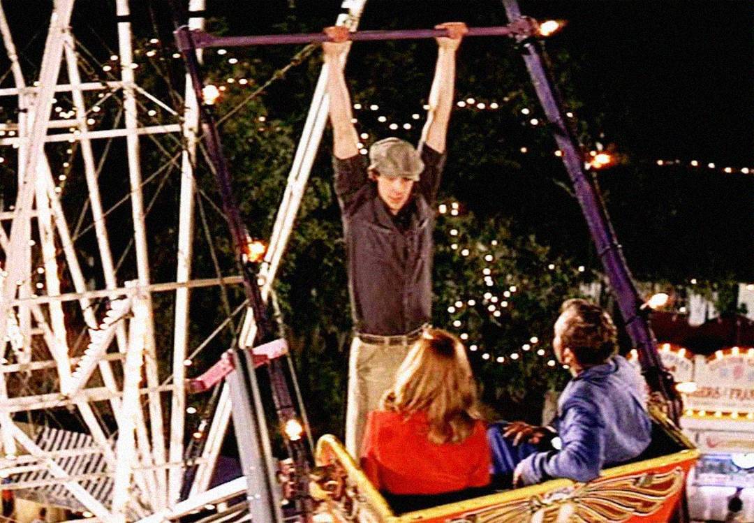 A young man hangs from the top of a Ferris wheel at night, looking down at a woman and man seated in a Ferris wheel car, with carnival lights glowing in the background.