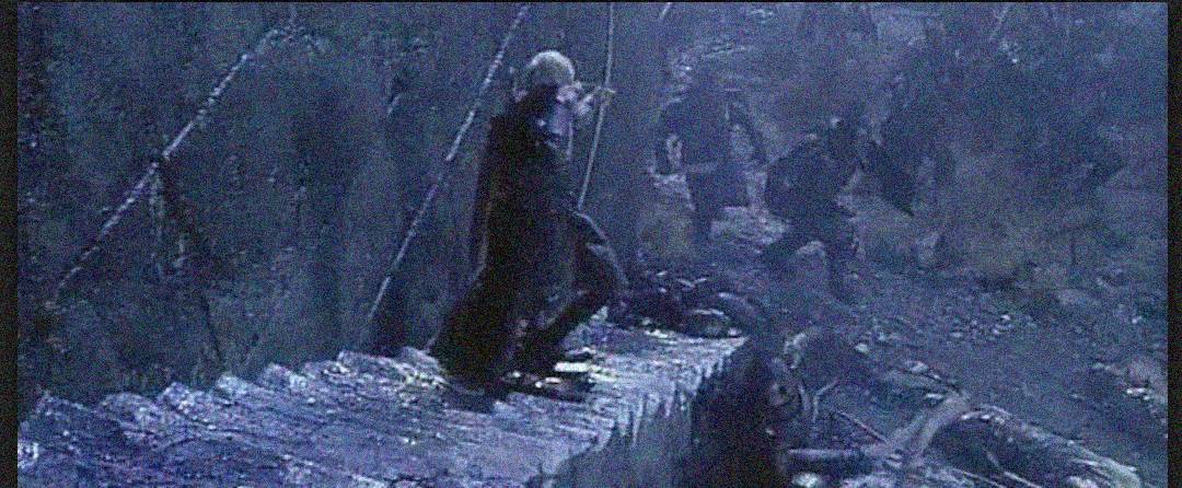 A group of people dressed in dark clothing are climbing and standing on a stone staircase at night, with shadowy figures and rough terrain in the background. The scene appears intense and action-filled.