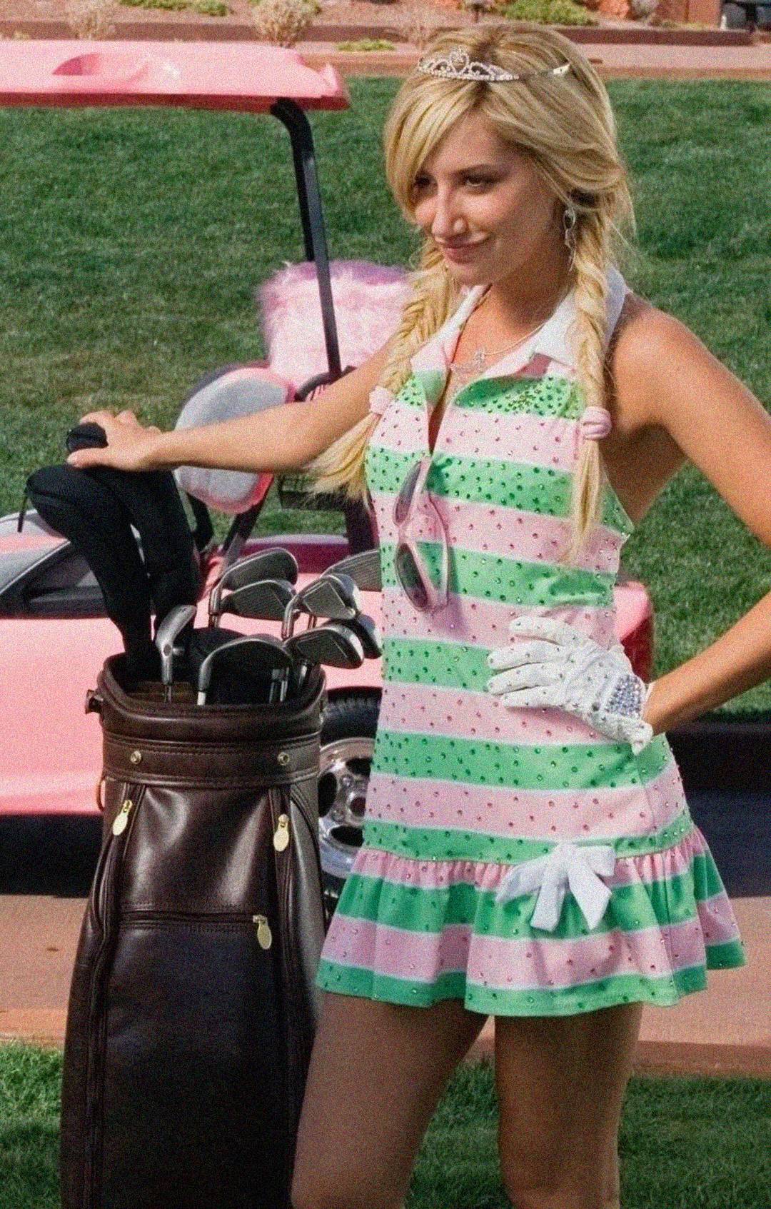 A woman wearing a pink and green striped dress, white gloves, a tiara, and two side braids stands by a golf bag with clubs. A pink golf cart and green grass are visible in the background.