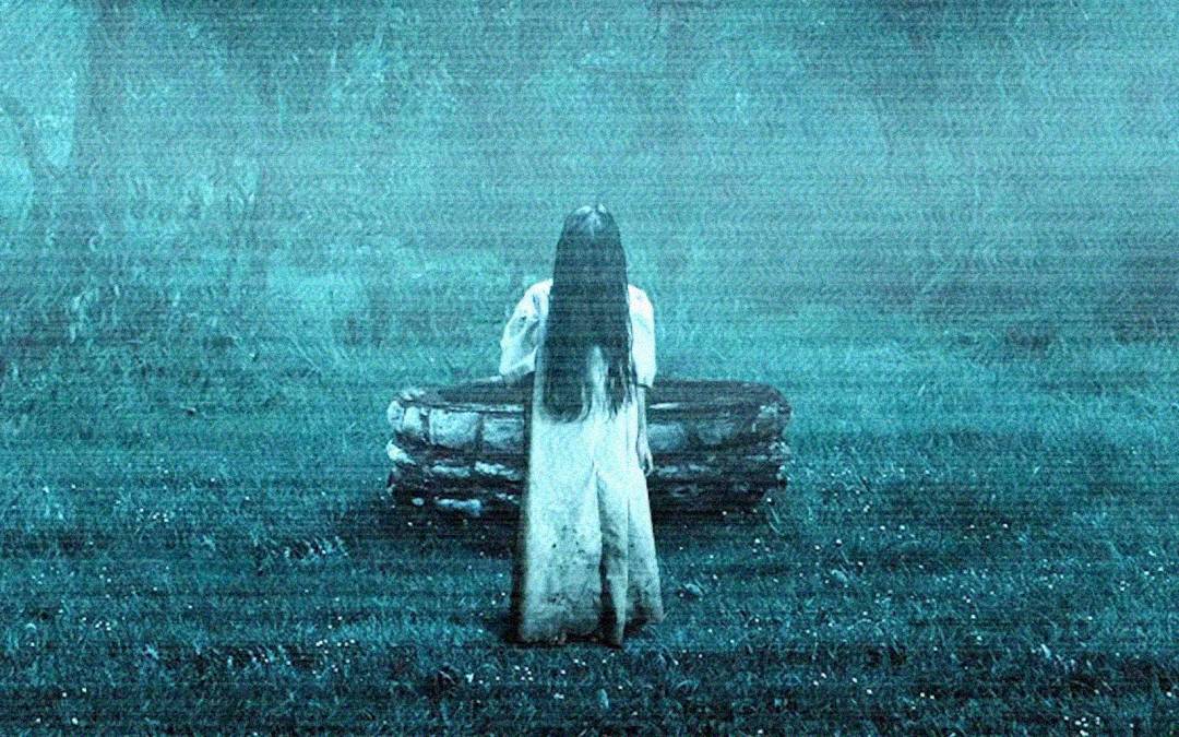 A figure with long black hair covering their face, wearing a white dress, stands in front of an old stone well in a misty, dimly lit grassy area. The scene has an eerie, supernatural atmosphere.