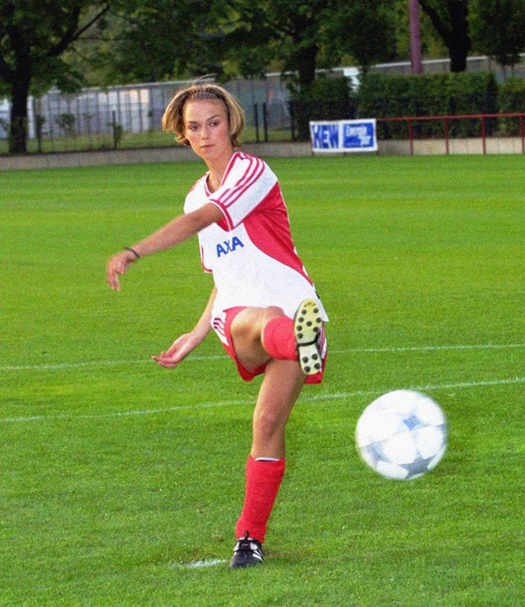 A young woman in a red and white soccer uniform is kicking a soccer ball on a green grass field, with trees and a fence visible in the background.