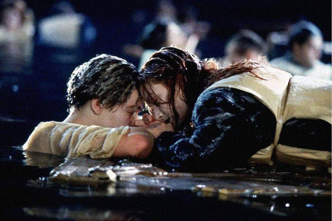 A young man and woman cling to a floating piece of debris in icy water at night, holding hands and looking at each other emotionally, surrounded by blurred figures in the background.