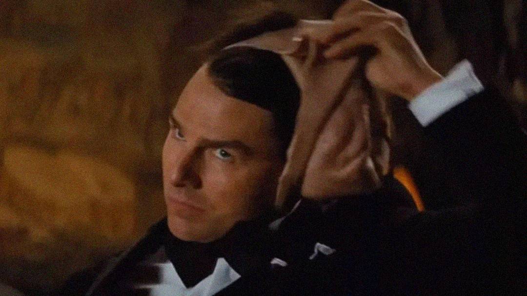 A person in formal attire pulls off a realistic-looking mask, revealing their true face underneath. The background is dimly lit and out of focus.