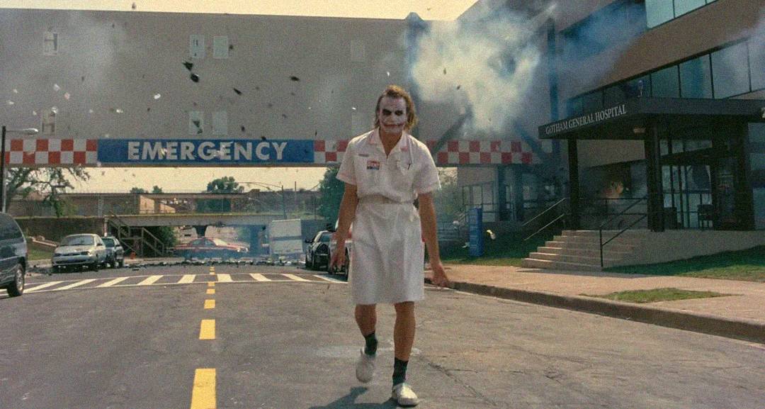 A person in a nurse costume with white face paint and smeared makeup walks away from a hospital with an "EMERGENCY" sign, as smoke and debris fill the air behind them from an explosion.