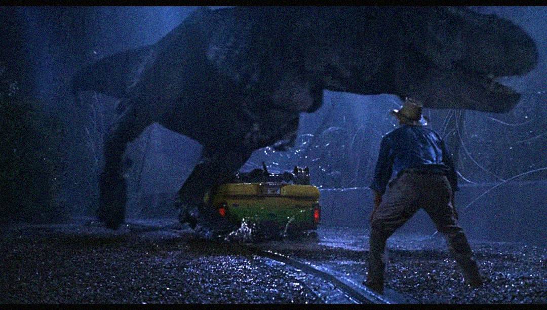 A large dinosaur towers over a man in a hat and raincoat standing near a green-and-yellow vehicle at night in the rain, illuminating a tense and dramatic scene.