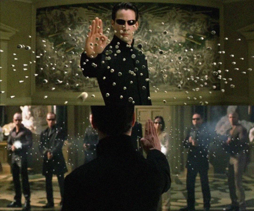 A man in black sunglasses and a long coat stands with his hand raised, stopping dozens of bullets mid-air. Armed people stand in the background, watching him in a dramatic, indoor setting.