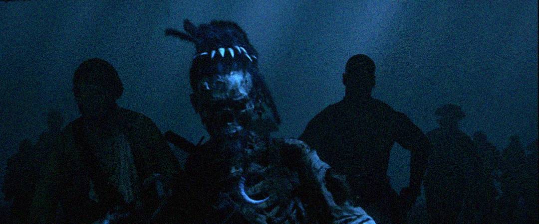 A decayed, skeletal zombie with tattered clothing and a pointed head is walking forward in a dark, foggy scene, surrounded by the shadowy figures of other zombies in the background.