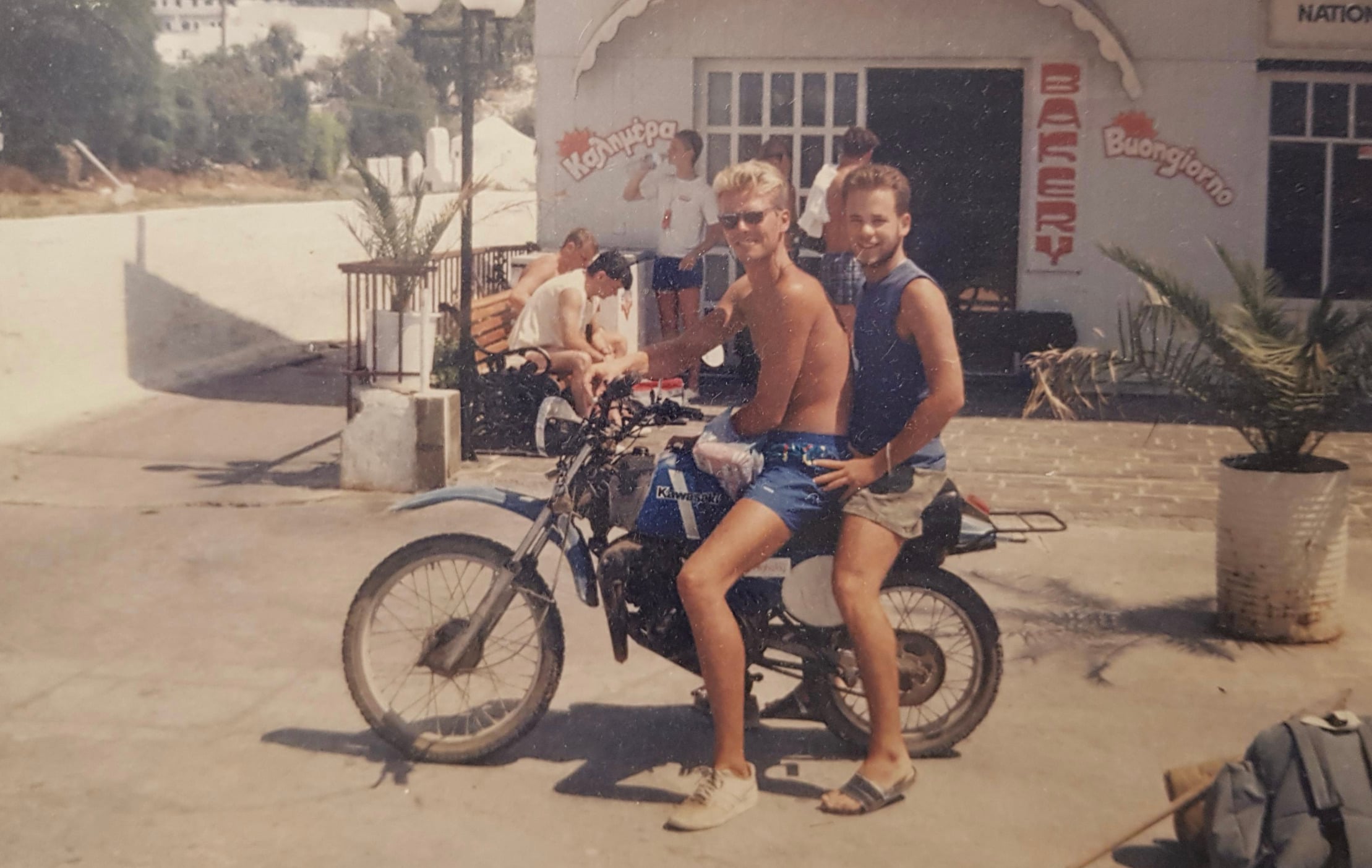Two people in summer clothes sit on a motorcycle, smiling at the camera. They are in front of a white building with bakery signs, potted plants, and other people relaxing nearby on a sunny day.