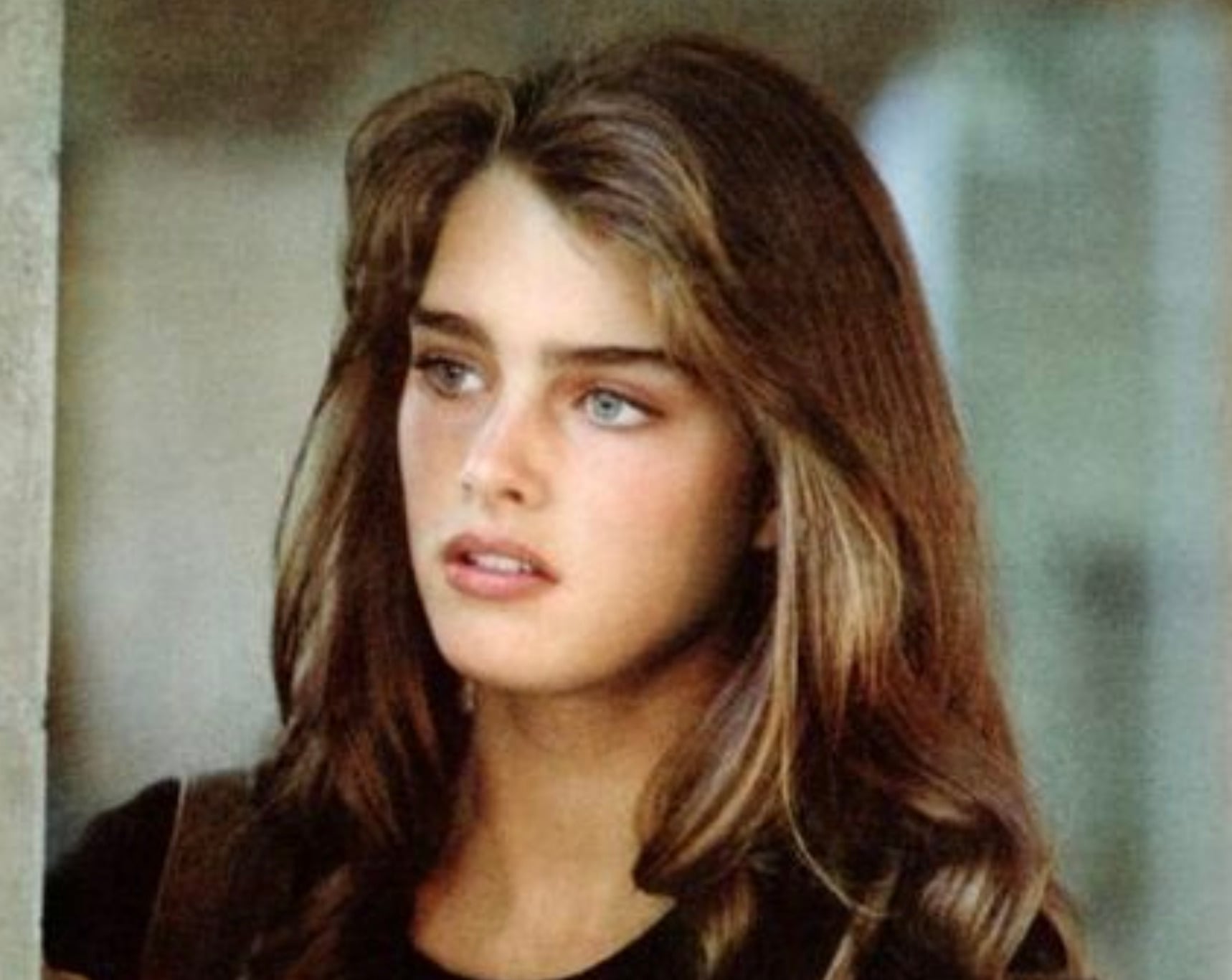 A young woman with long wavy brown hair and blue eyes gazes slightly to the side with a serious expression, wearing a dark top. The background is softly blurred.