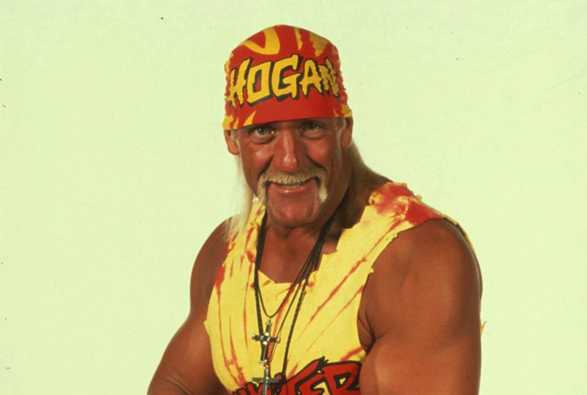 A muscular man wearing a red and yellow bandana, a yellow sleeveless shirt, and several necklaces, posing with a confident smile against a plain light background.