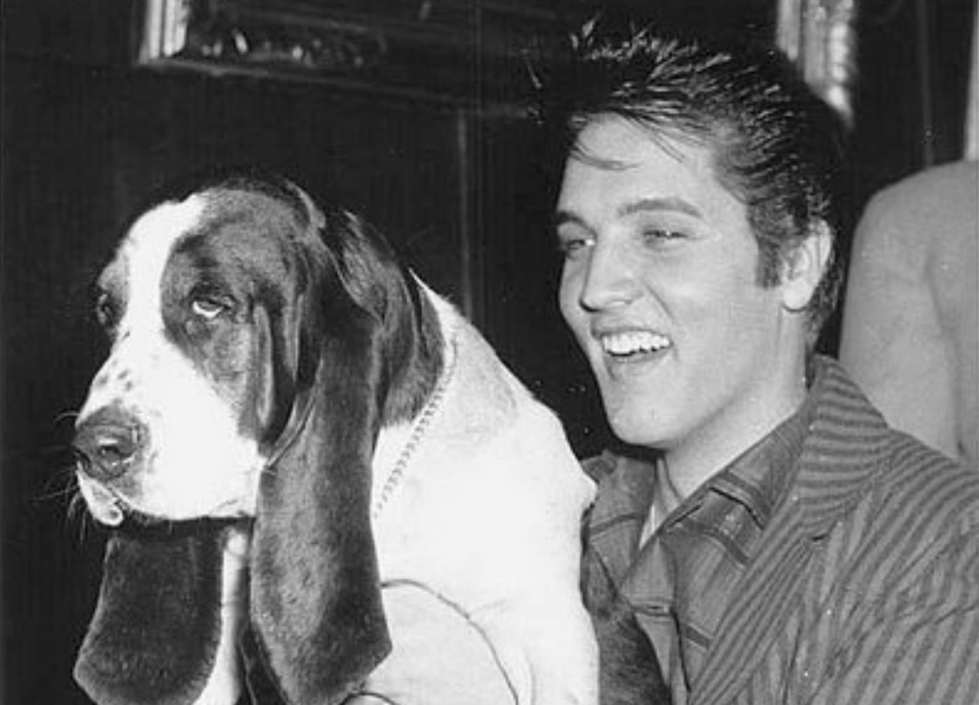 A young man with dark hair smiles while holding a large, floppy-eared dog. The photo is black and white, and the man is wearing a striped jacket. The dog looks relaxed with its head turned slightly to the side.