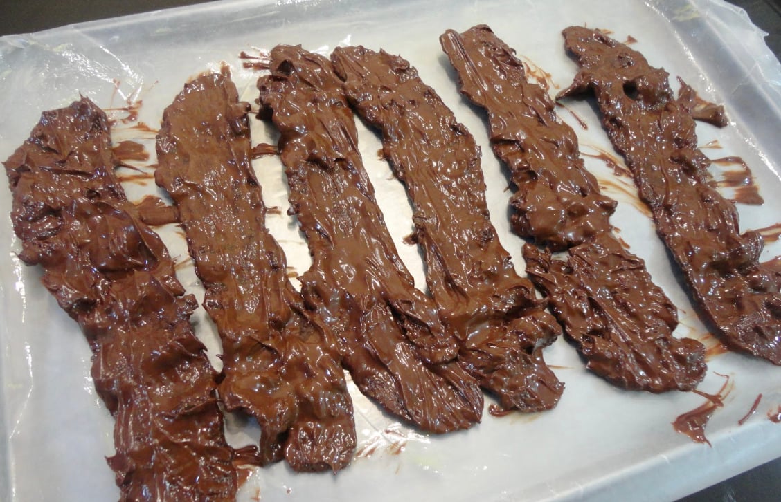 Six strips of bacon coated in melted chocolate are laid out on a sheet of wax paper to cool and harden.