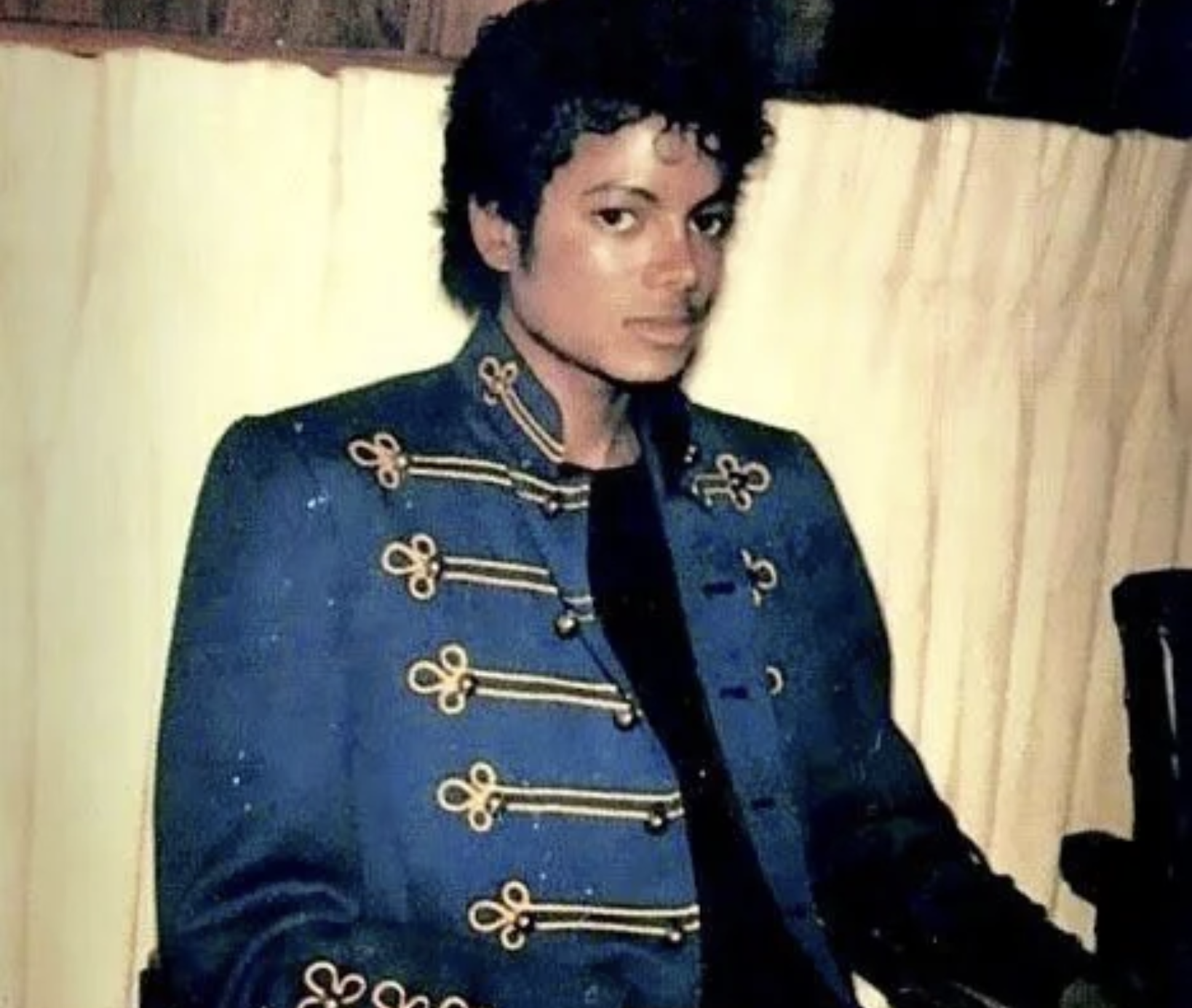 A person with curly hair wearing a dark blue military-style jacket with gold detailing sits indoors against a light-colored curtain, looking towards the camera with a neutral expression.